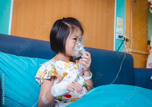Sad asian child holds a mask vapor inhaler for treatment of asthma. Breathing through a steam nebulizer.Illness girl admitted in hospital while saline intravenous (IV) on hand.
