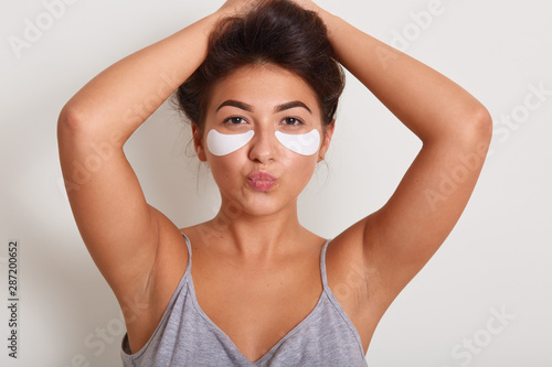 Studio shot of young beautiful woman with clean perfect skin with cosmetic patches on face. Beauty portrait of charming girl keeps hands up and lips rounded, looks at camera, isolated over white wall.