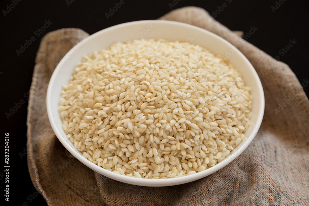 Organic Arborio rice in a white bowl on a black background, low angle view. Close-up. Organic Arborio rice in a white bowl on a black background, low angle view. Close-up.