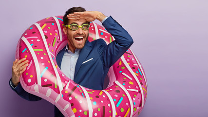 Photo of overjoyed male office worker focused into distance, keeps palm near forehead, pretends being on beach, wears goggles for swimming, poses in inflated swimring, notices something funny