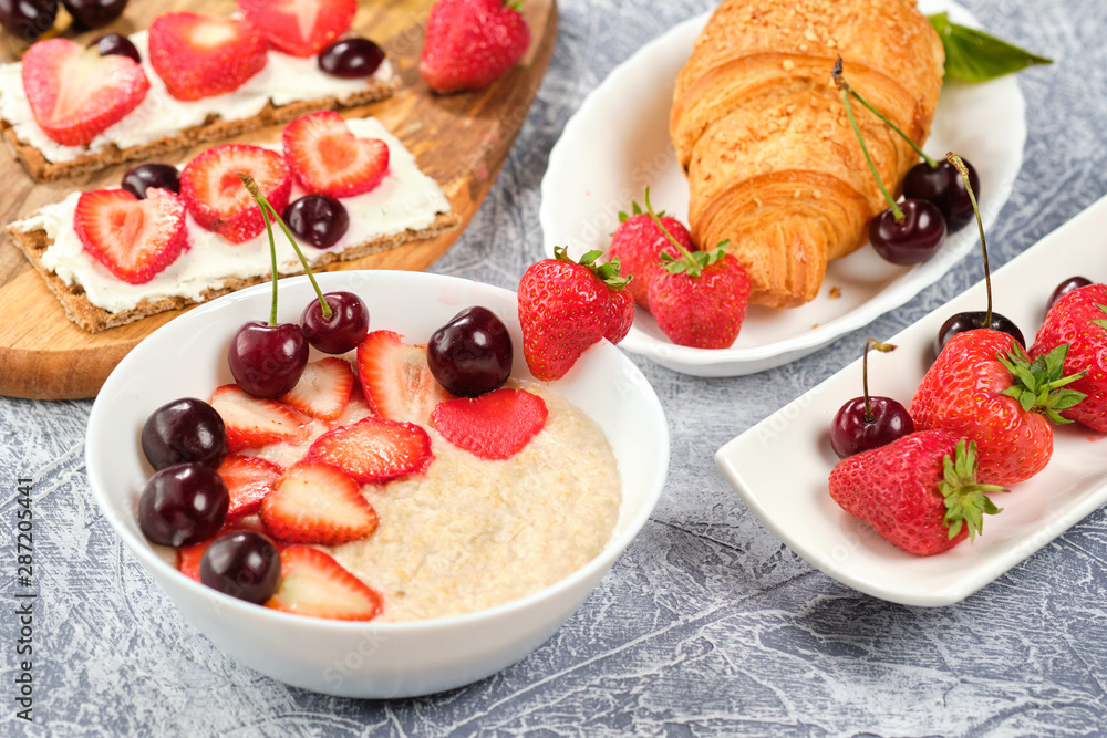white bowl of porridge with strawberries and cherries, croissants and crispbread with cheese and strawberries on a gray background white bowl of porridge with strawberries and cherries, croissants and crispbread with cheese and strawberries on a gray background