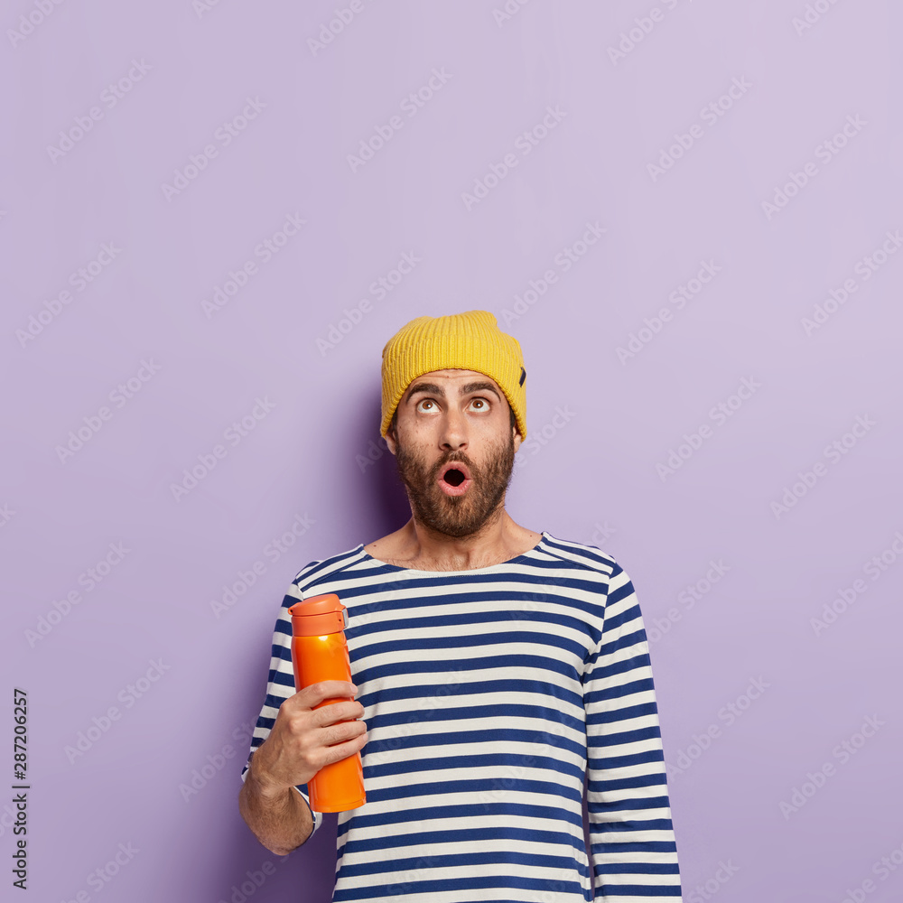 Photo of astonished young male model focused upwards, holds orange thermos, wears striped jumper and yellow hat, has surprised expression, stands against purple studio wall, free space above Photo of astonished young male model focused upwards, holds orange thermos, wears striped jumper and yellow hat, has surprised expression, stands against purple studio wall, free space above