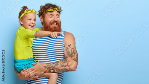 Happy surprised redhaired bearded man stands with daughter, look glafully into distance, wear protective goggles for swimming, isolated over blue background. Childhood and fatherhood concept