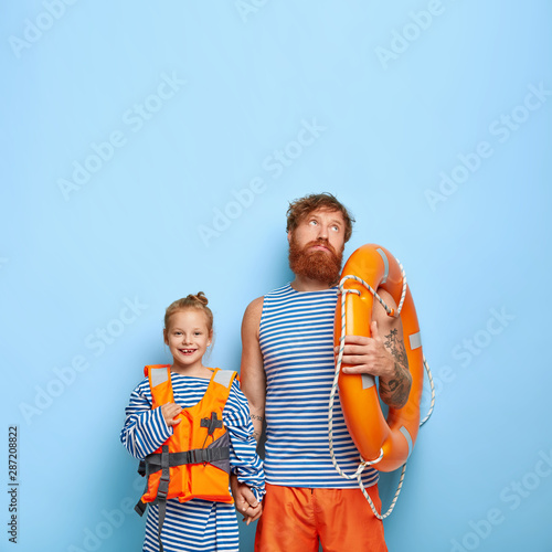 Cheerful smiling daughter wears oversized sailor jumper and inflated lifejacket, holds hand of dad who has indifferent sad expression, carries lifebuoy, feels tired of teaching child to swim