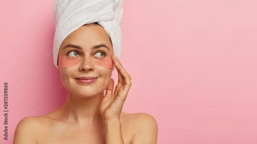 Studio shot of good looking satisfied woman with beauty pads under eyes, has daily spa procedures, healthy fresh skin, looks with green eyes aside, stands against pink background, free space aside Studio shot of good looking satisfied woman with beauty pads under eyes, has daily spa procedures, healthy fresh skin, looks with green eyes aside, stands against pink background, free space aside