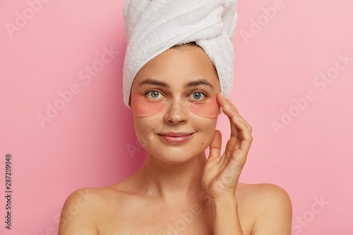 Close up shot of beautiful European female model makes spa procedures after taking shower, applies collagen patches under eyes, has anti aging treatment, stands indoor, isolated on pink studio wall