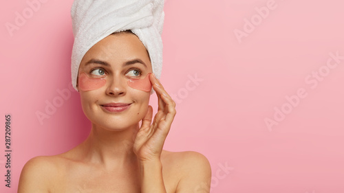 Studio shot of good looking satisfied woman with beauty pads under eyes, has daily spa procedures, healthy fresh skin, looks with green eyes aside, stands against pink background, free space aside photo