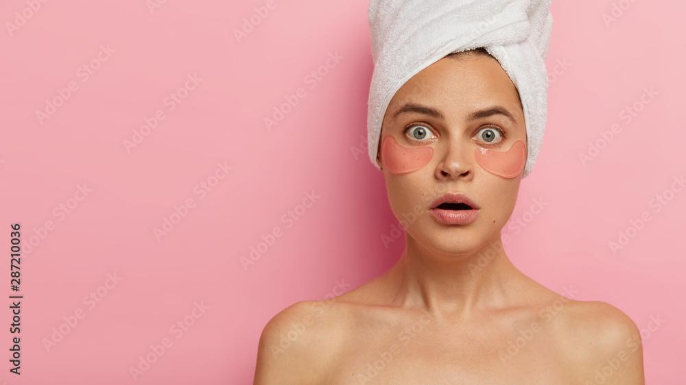 Image of shocked attractive young woman has bated breath, natural makeup, wears hydrogel pads under eyes, removes wrinkles, has rejuvenation treatment, stands topless against pink studio wall. Image of shocked attractive young woman has bated breath, natural makeup, wears hydrogel pads under eyes, removes wrinkles, has rejuvenation treatment, stands topless against pink studio wall.