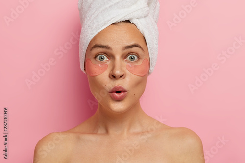 Under eye treatment and body care concept. Shocked young Caucasian woman applies beauty patches, removes dark circles and puffiness, looks surprisingly at herself in mirror, stands topless indoor