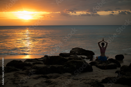 Woman sitting on the rock, practicing yoga and enjoying ocean view. View from back. Hands in namaste mudra. Yoga at the beach.