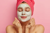 Headshot of lovely girl keeps clenched fists near face, wears facial cleansing mask, keeps eyes shut, has bare shoulders, healthy perfect skin, cares about her body, isolated on pink wall. Wellness