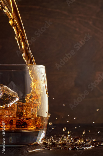 A glass of whiskey with ice on the wooden table. Top in a glass filled with whiskey. Right splashes from the drink. photo