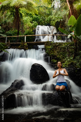Healthy life. Yoga retreat. Young woman sitting on the rock, meditating, practicing yoga, pranayama with namaste mudra at waterfall. Slow shutter speed, motion photography. Jembong waterfall, Bali © Olga