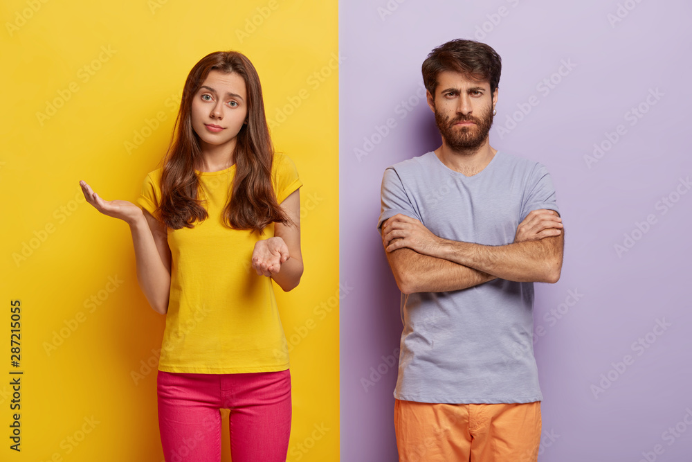 Hesitant young woman spreads hands with doubt, wears casual clothes, dissatisfied man keeps hands crossed, discontent with something, stands against two colored studio wall. Negative emotions Hesitant young woman spreads hands with doubt, wears casual clothes, dissatisfied man keeps hands crossed, discontent with something, stands against two colored studio wall. Negative emotions