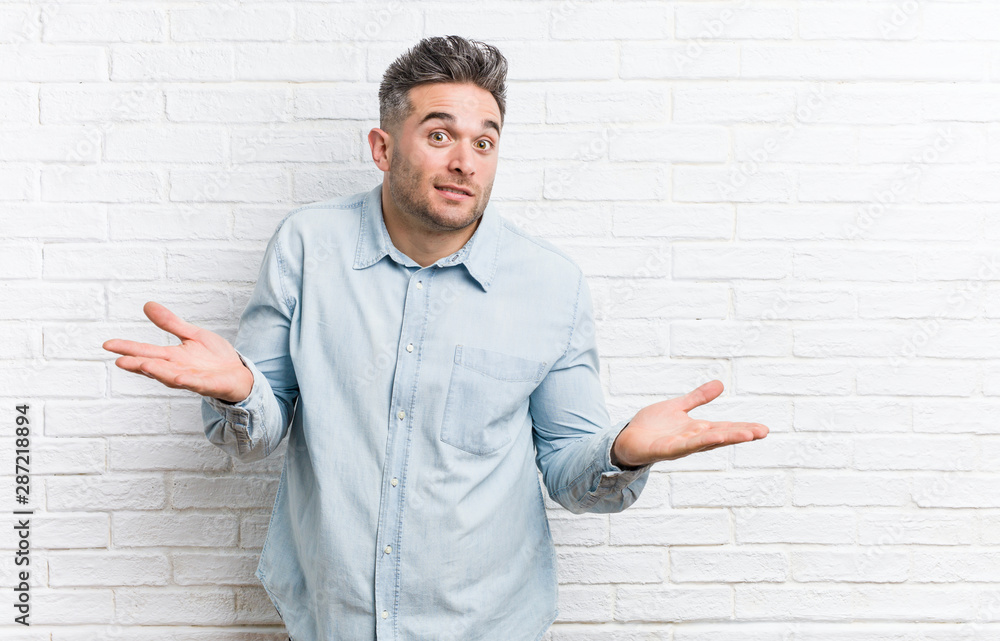 Young handsome man against a bricks wall doubting and shrugging shoulders in questioning gesture. Young handsome man against a bricks wall doubting and shrugging shoulders in questioning gesture.
