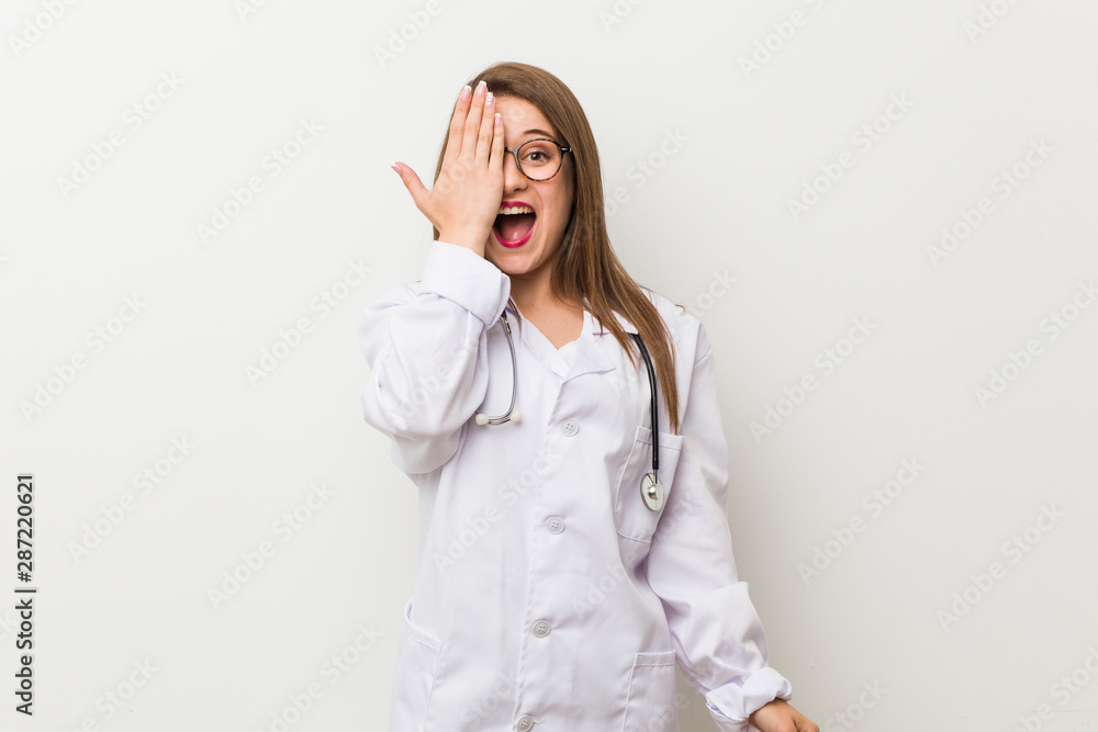 Young doctor woman against a white wall having fun covering half of face with palm.