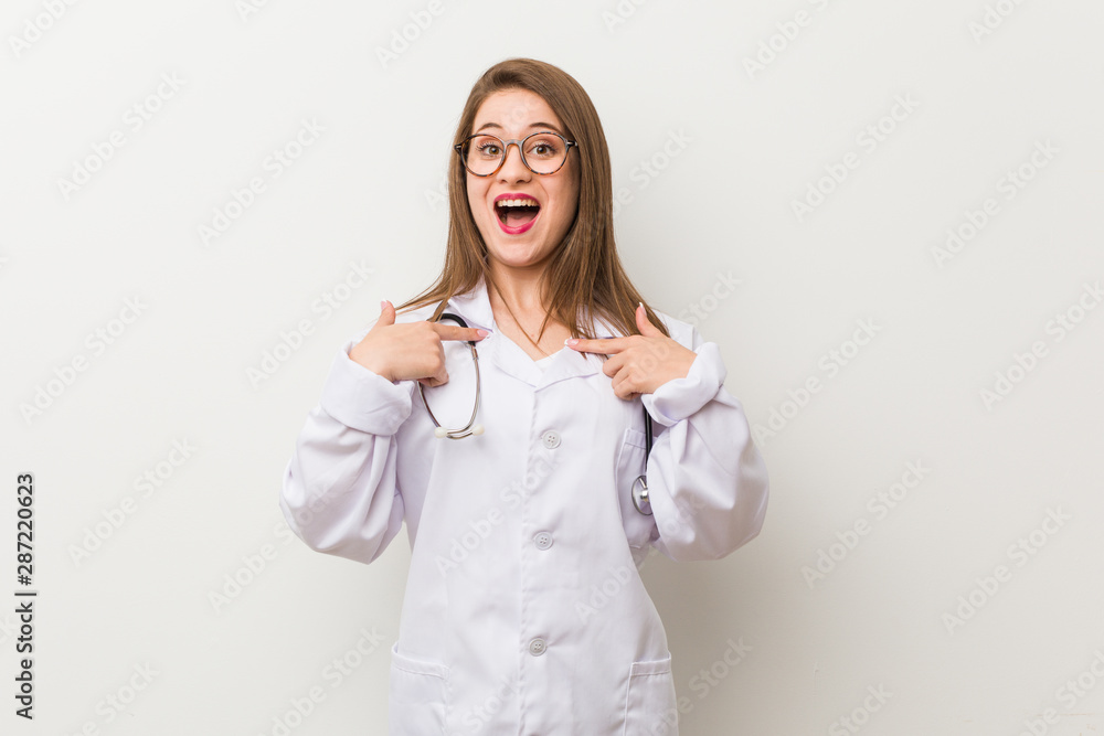 Young doctor woman against a white wall surprised pointing with finger, smiling broadly. Young doctor woman against a white wall surprised pointing with finger, smiling broadly.