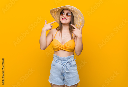 Young caucasian woman wearing a straw hat, summer look smiles, pointing fingers at mouth.