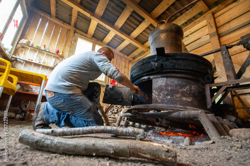 Man feeding wood in the hot oven of an traditional craft installation for making alcoholic beverage in a house in Maramures, Romania