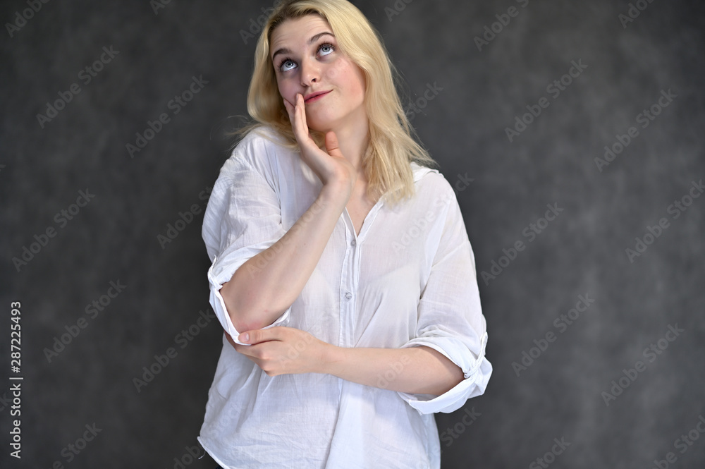 Portrait of a cute blonde girl, a young woman in a white shirt with beautiful curly hair on a gray background. Shows hands to the sides. Beauty, brightness, emotions. Portrait of a cute blonde girl, a young woman in a white shirt with beautiful curly hair on a gray background. Shows hands to the sides. Beauty, brightness, emotions.