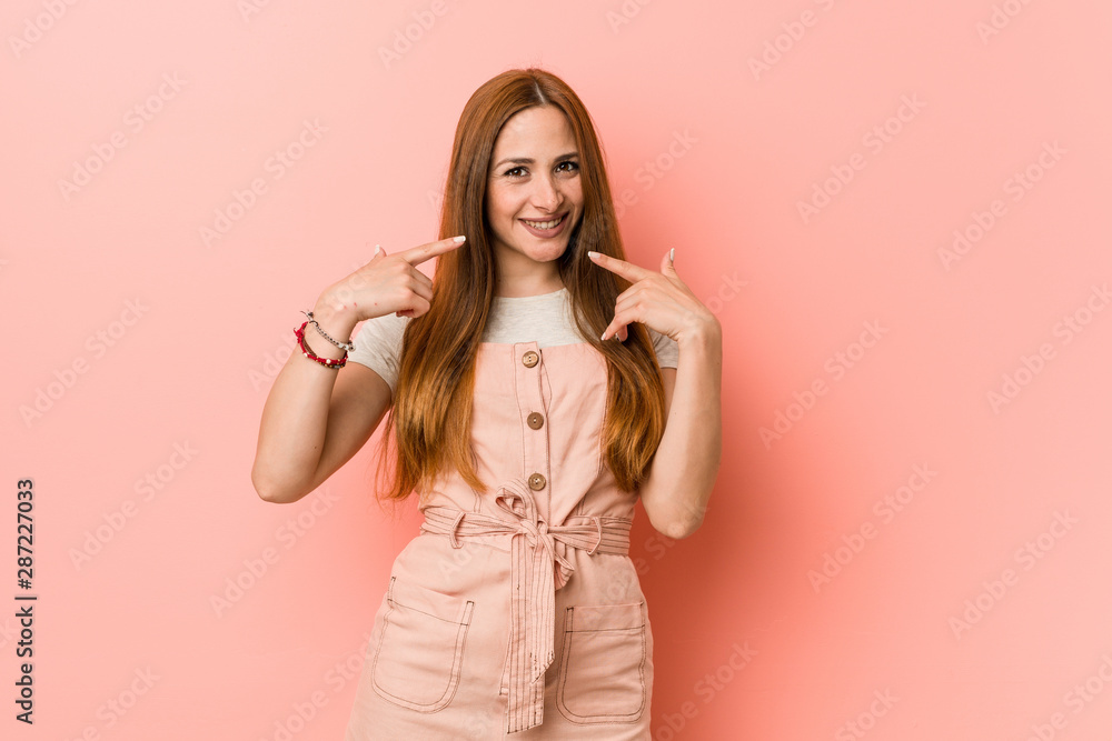 Young ginger woman with freckles smiles, pointing fingers at mouth. Young ginger woman with freckles smiles, pointing fingers at mouth.