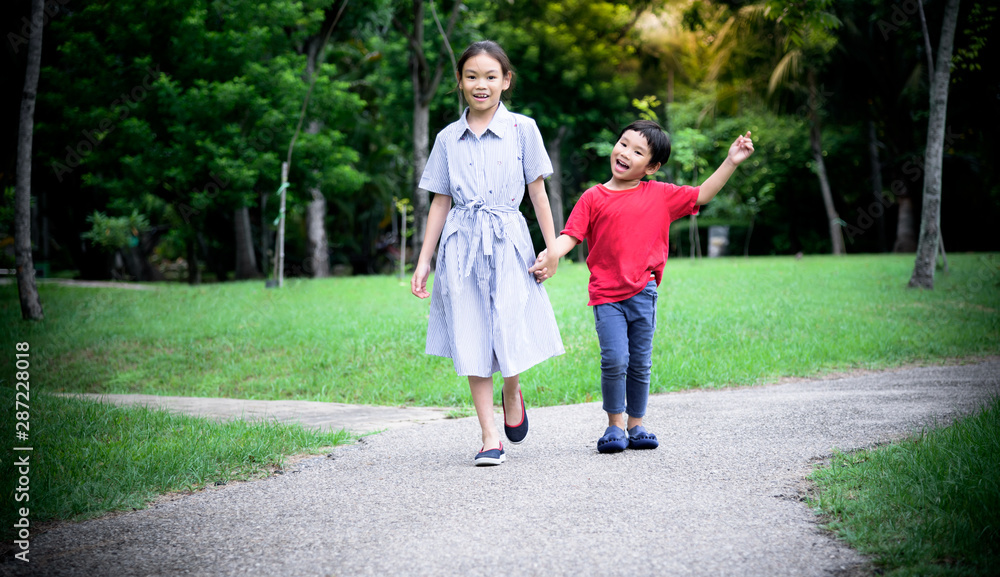 Asian children, sisters and brothers held hands walked on the streets in the park Are smiling, fun and happy with blurred soft of green trees background, to children and family concept.