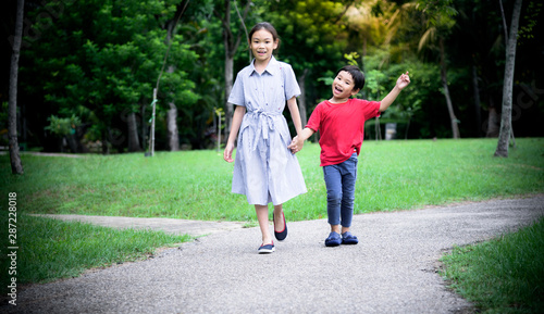 Fototapeta Naklejka Na Ścianę i Meble -  Asian children, sisters and brothers held hands walked on the streets in the park Are smiling, fun and happy with blurred soft of green trees background, to children and family concept.