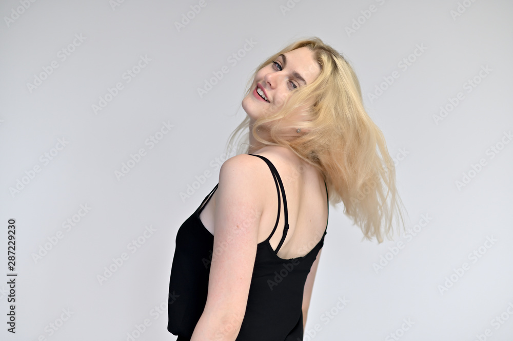 Fototapeta premium Portrait of a cute blonde student girl, a young woman in a black dress with beautiful curly hair on a white background in different poses. Shows hands to the sides. Beauty, brightness, emotions.