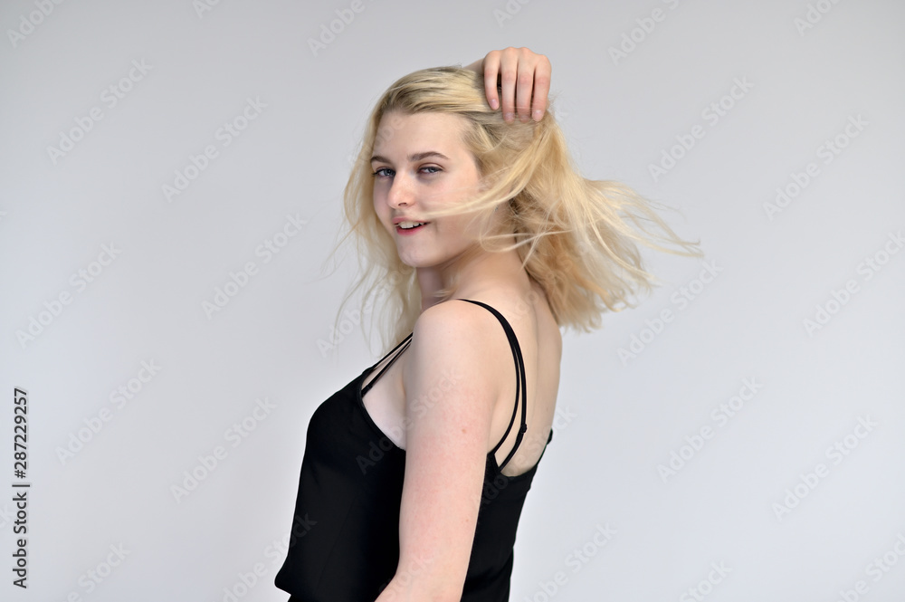 Fototapeta premium Portrait of a cute blonde student girl, a young woman in a black dress with beautiful curly hair on a white background in different poses. Shows hands to the sides. Beauty, brightness, emotions.