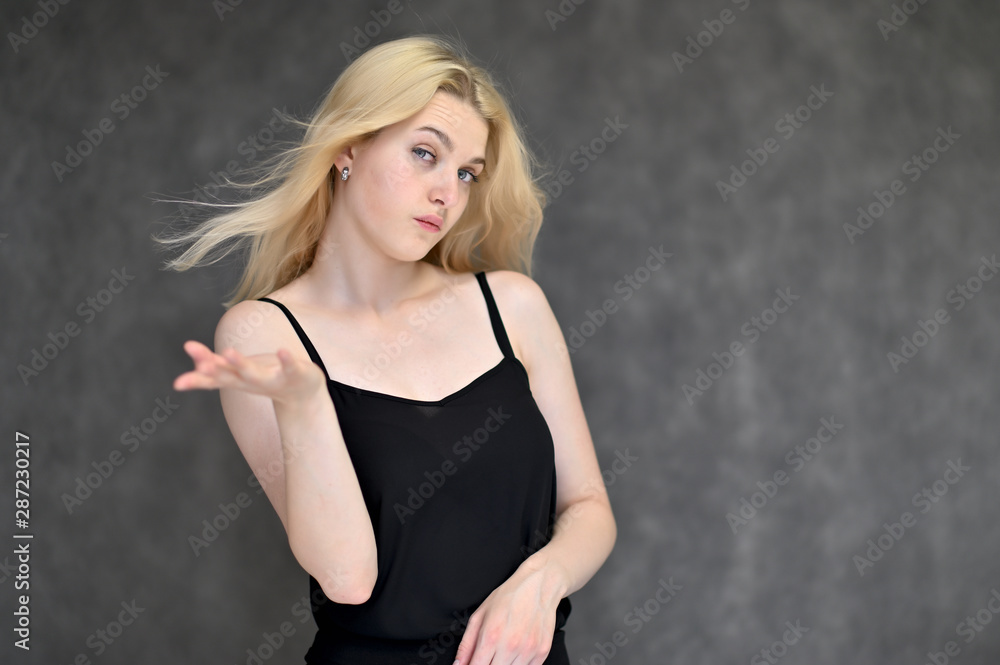 Portrait of a cute blonde student girl, a young woman in a black dress with beautiful curly hair on a gray background in different poses. Shows hands to the sides. Beauty, brightness, emotions. Portrait of a cute blonde student girl, a young woman in a black dress with beautiful curly hair on a gray background in different poses. Shows hands to the sides. Beauty, brightness, emotions.