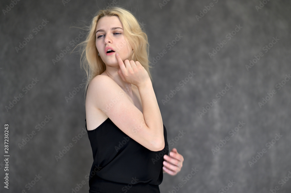 Portrait of a cute blonde student girl, a young woman in a black dress with beautiful curly hair on a gray background in different poses. Shows hands to the sides. Beauty, brightness, emotions. Portrait of a cute blonde student girl, a young woman in a black dress with beautiful curly hair on a gray background in different poses. Shows hands to the sides. Beauty, brightness, emotions.
