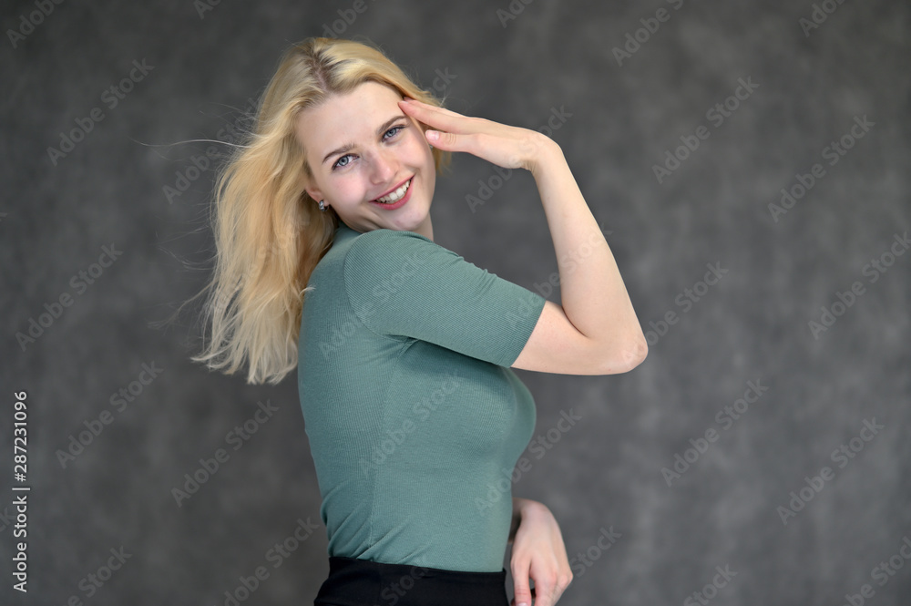 Portrait of a cute blonde student girl, a young woman in a green T-shirt with beautiful curly hair on a gray background in different poses. Shows hands to the sides. Beauty, brightness, emotions. Portrait of a cute blonde student girl, a young woman in a green T-shirt with beautiful curly hair on a gray background in different poses. Shows hands to the sides. Beauty, brightness, emotions.