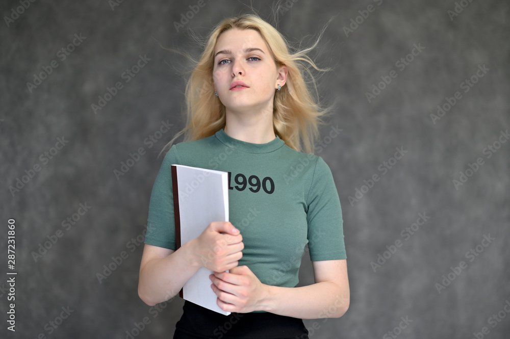 Portrait of a cute blonde student girl, a young woman in a green T-shirt with beautiful curly hair on a gray background in different poses. With a folder in his hands. Beauty, brightness, emotions.