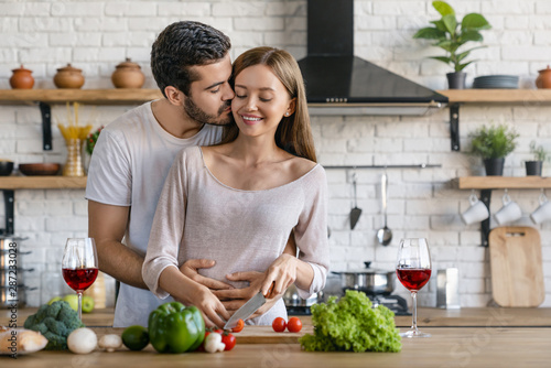 Beautiful cheerful young couple cooking dinner while man kissing his girlfriend standing in the kitchen at home