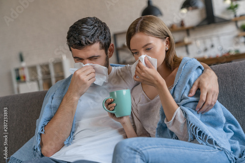 Cold and flu season. Young couple sitting on sofa covered with blanket, sneezing and blowing nose