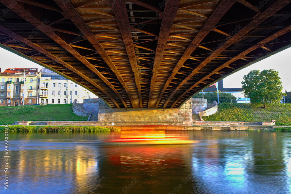 Long exposure image of a small boat passing through the Neris River under the Green Bridge in Vilnius, Lithuania.