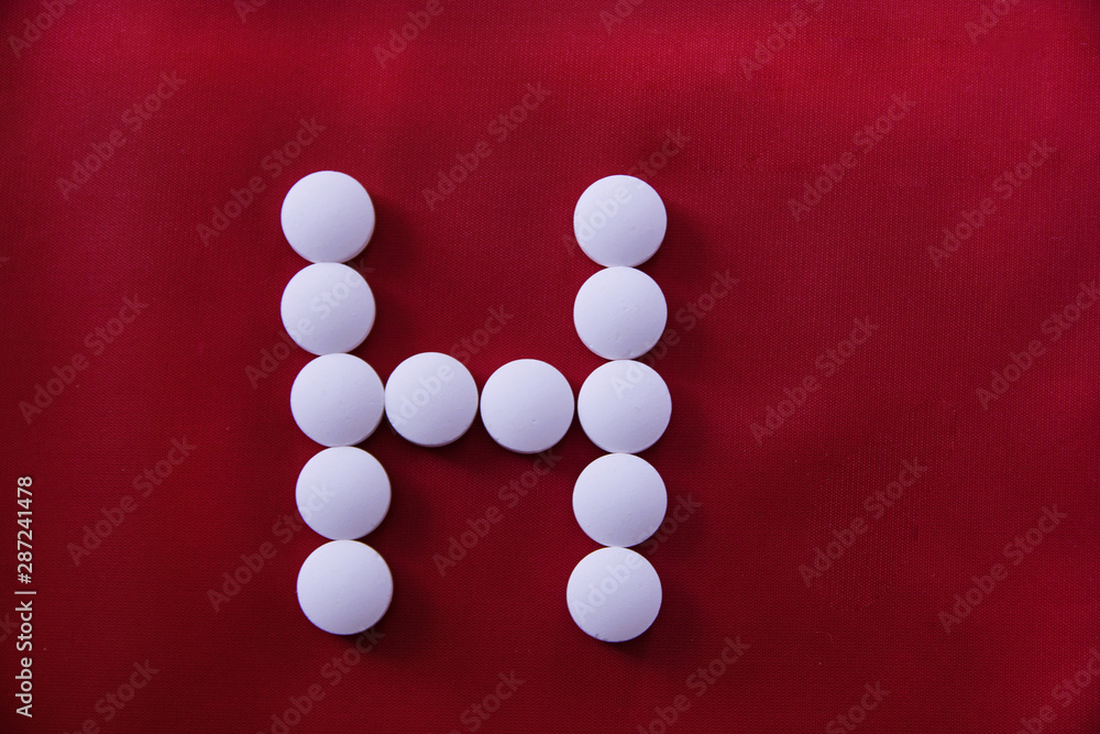 White round medical pills lie on a red background in the form of a letter H White round medical pills lie on a red background in the form of a letter H