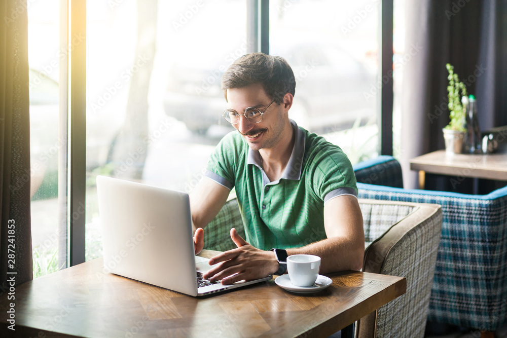 Young happy successful businessman with mustache in green t-shirt sitting and working on laptop with toothy smile. business and freelancing concept. indoor shot near big window at daytime.