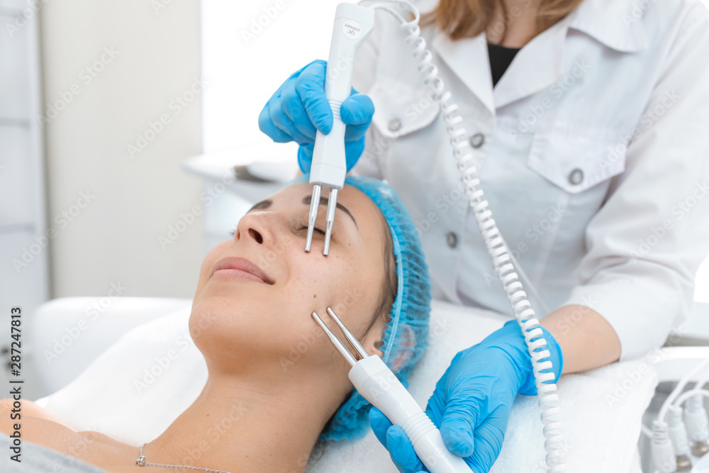 Naklejka premium Beautician procedure of microcurrent therapy of the patient's face. A young girl is undergoing a course of spa treatments in the office of a beautician. Moisturizing, cleaning and facial skin care. 