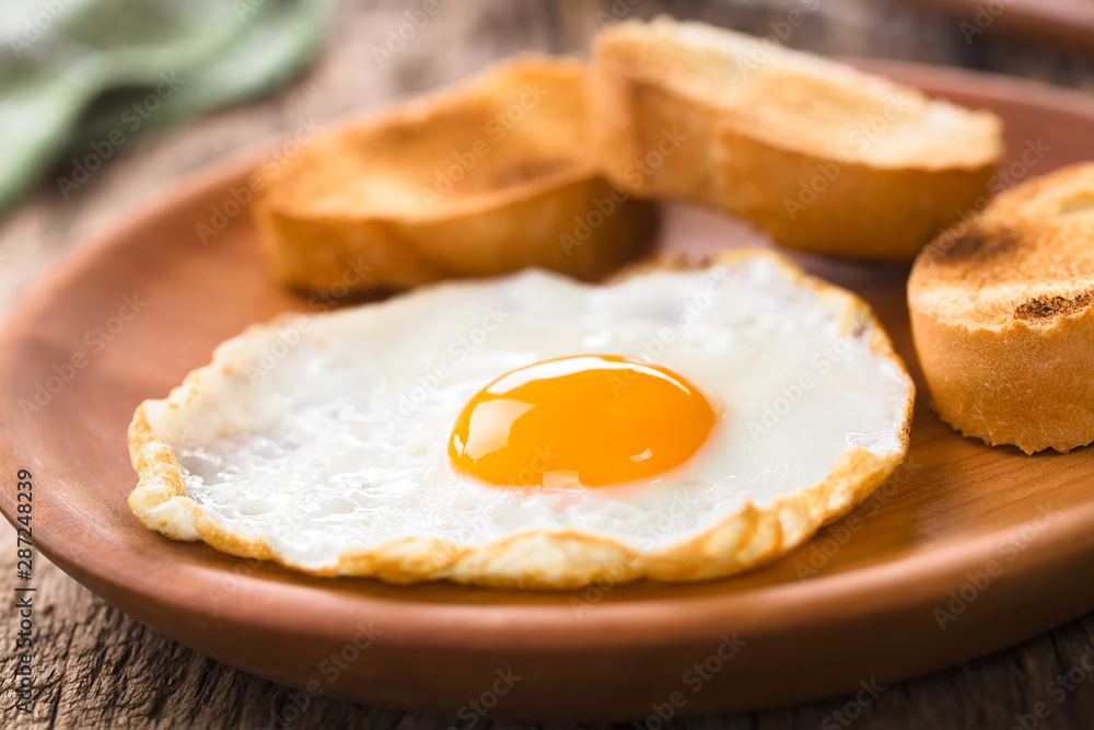 One fresh fried egg sunny side up with toasted baguette slices on the side served on wooden plate (Selective Focus, Focus on the front of the egg yolk) One fresh fried egg sunny side up with toasted baguette slices on the side served on wooden plate (Selective Focus, Focus on the front of the egg yolk)