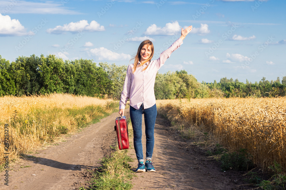 Beautiful young woman with a suitcase on the road between wheat fields in the background. Travel concept, vacation