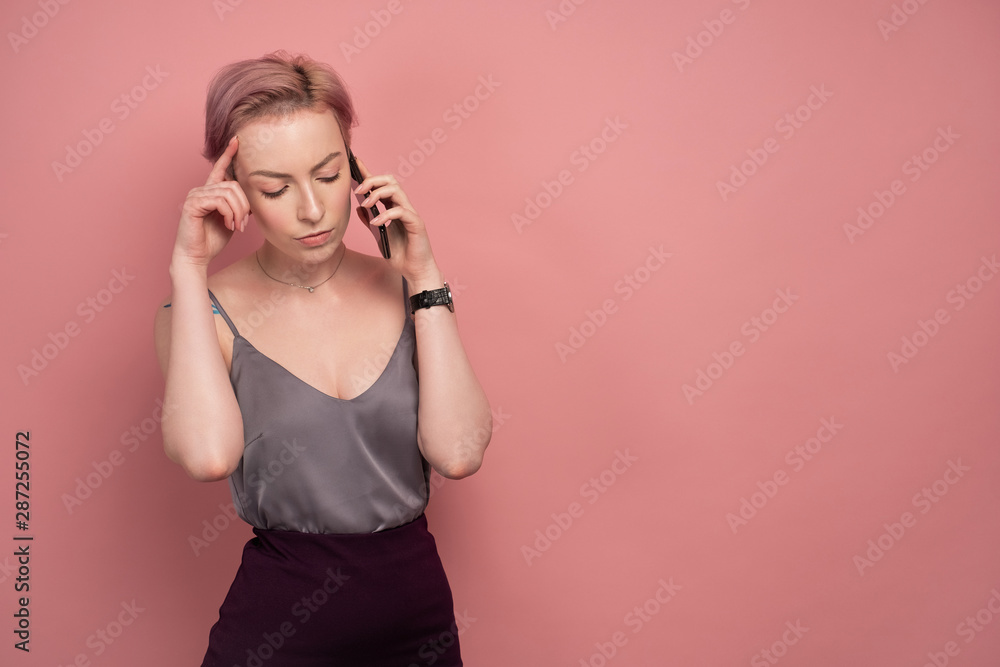 A girl with short pink hair and a tattoo in a gray top and speaks on the phone, listening carefully, on a pink background. A girl with short pink hair and a tattoo in a gray top and speaks on the phone, listening carefully, on a pink background.