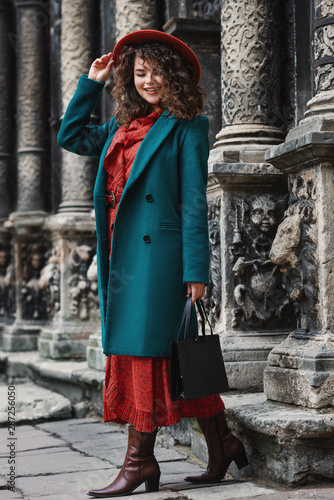 Outdoor full-length portrait of happy smiling fashionable lady wearing trendy orange dress, hat, green classic coat, brown cowboy ankle boots, holding black handbag, walking in street of European city
