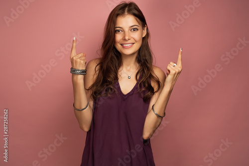 The brunette is standing on a pink background smiling, looking at the camera with her fingers pointing to the top.