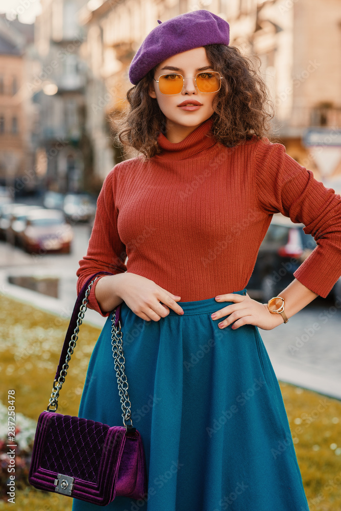 Outdoor autumn portrait of young beautiful elegant lady wearing wrist watch, purple beret, orange sunglasses, turtleneck, blue skirt, holding quilted velour bag, posing in street of European city