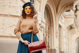Outdoor autumn fashion portrait of elegant lady wearing trendy leather beret, beige turtleneck, skirt, orange sunglasses, wrist watch, holding red bag, posing in street of European city. Copy space