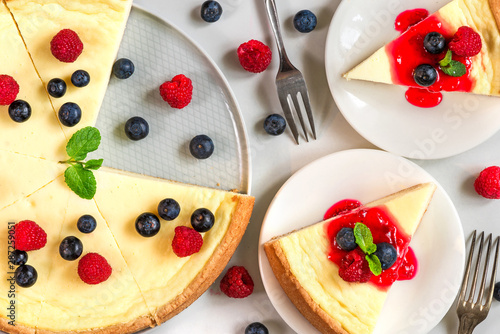 classic New York cheesecake with two slices of cake in plates with fresh berries, jam and mint on white marble table