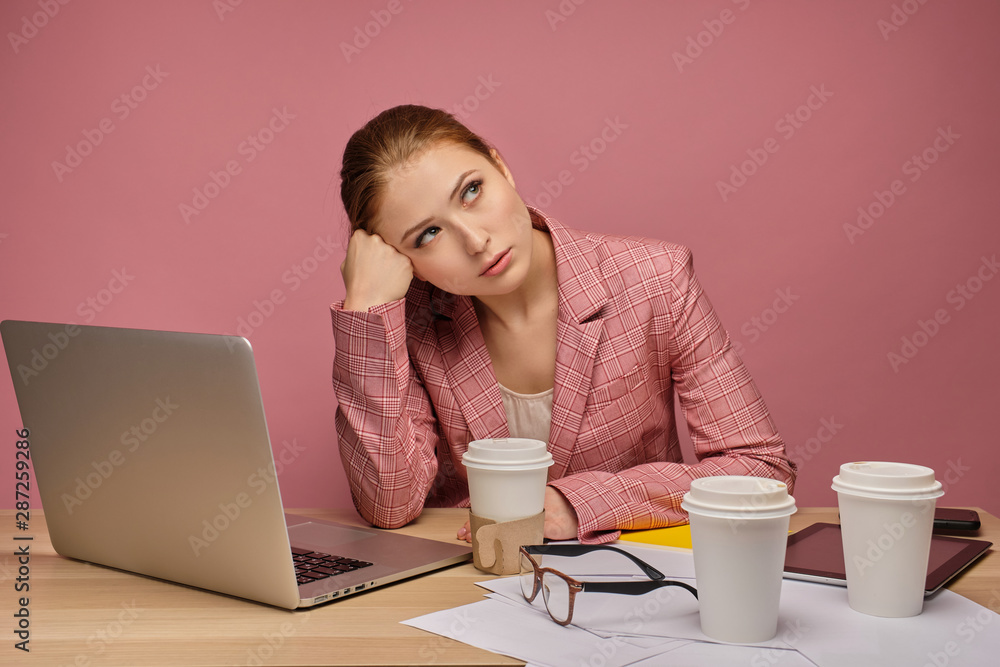 Redhead girl in a jacket sitting at a table in front of a laptop on a pink background, with head on hand and looking upwardly. Redhead girl in a jacket sitting at a table in front of a laptop on a pink background, with head on hand and looking upwardly.