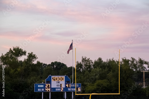 Sunset view of football end zone with goal posts, American flag flying and score board during evening at local high school
