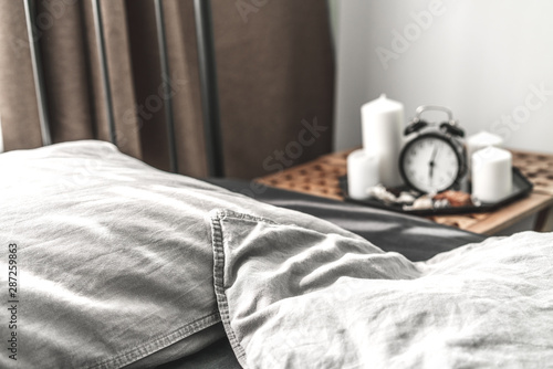 Cozy gray bedroom interior with comfortable bed near the wooden night table with candle, alarm clock and dried flowers. Cozy atmosphere at home. Warming up autumn evenings. Vintage furniture.